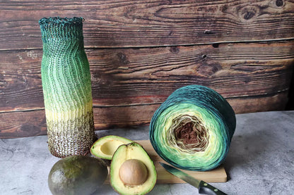 Auf dem Bild ist vor einer Holzwand der Bobbel "Avocado" mit einem dazugehörigen Strickmuster zu sehen. Als Dekoration sind geschlossene und aufgeschnittene Avocados und ein Schneidebrett mit Messer dabei.